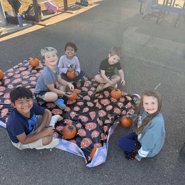 Five children sit on a blanket with pumpkins on a paved surface; two children hold pumpkins.