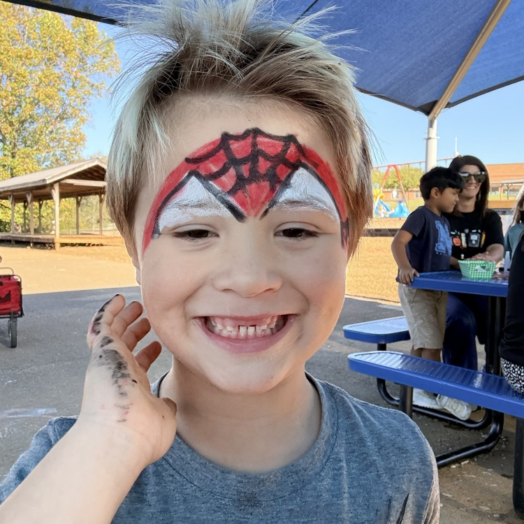 A child with Spider-Man face paint smiles with one hand raised under a blue canopy.