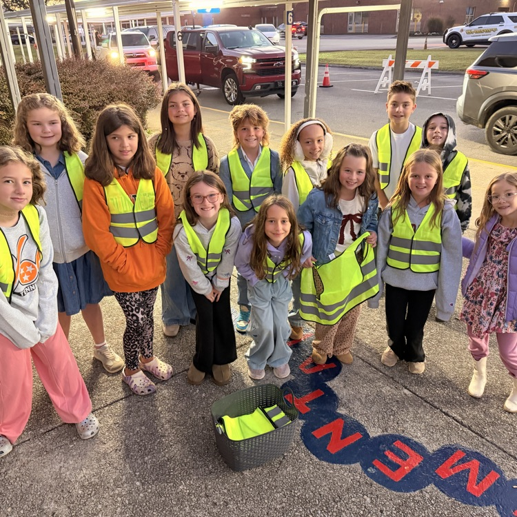 Children wearing yellow safety vests standing on the sidewalk. One child holds a bag. Letters spell out word.