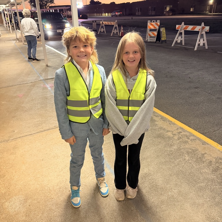Two children in high-visibility vests stand next to each other on a sidewalk near traffic barriers.