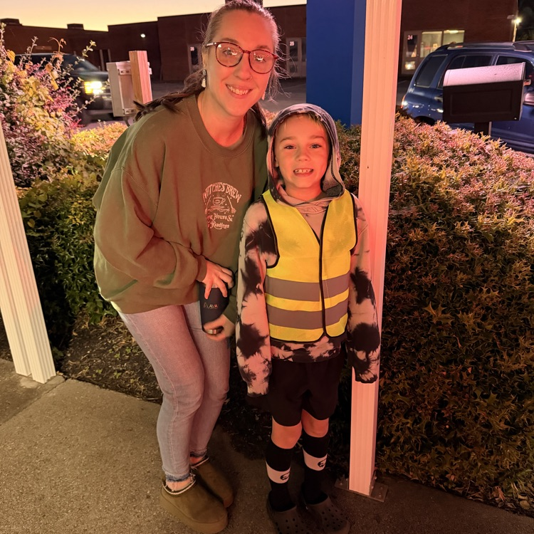 Woman and child in reflective vests pose outside with shrubs and parked cars in background.