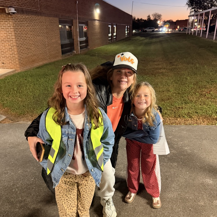 Adult and two children pose on a sidewalk outside a brick building. Background includes a grassy field.