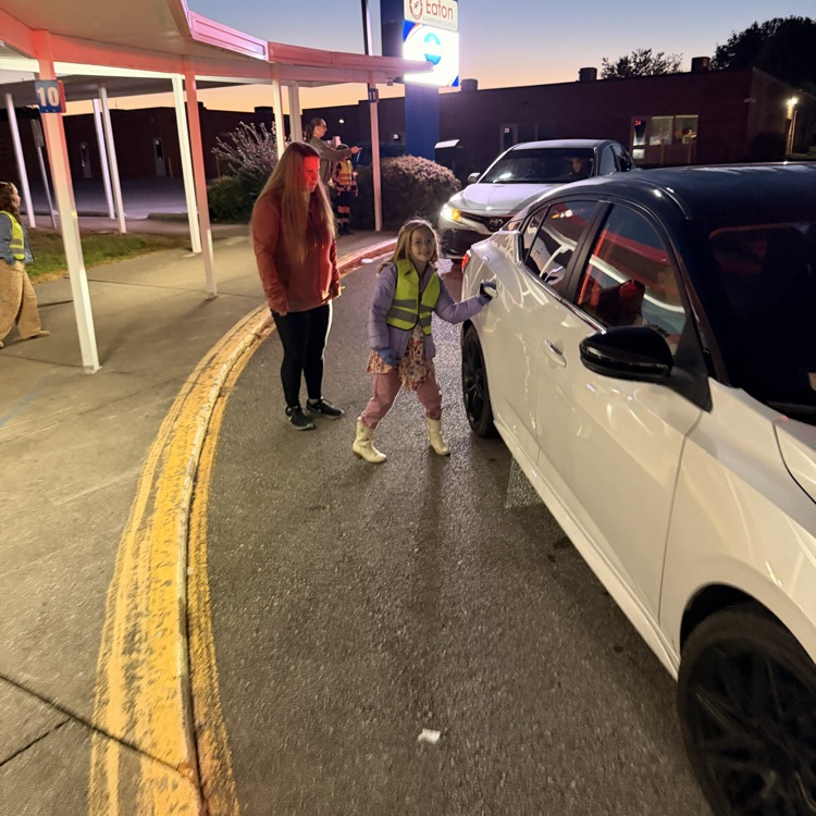 Two women, one in reflective gear, stand near a white car, with one opening a door.