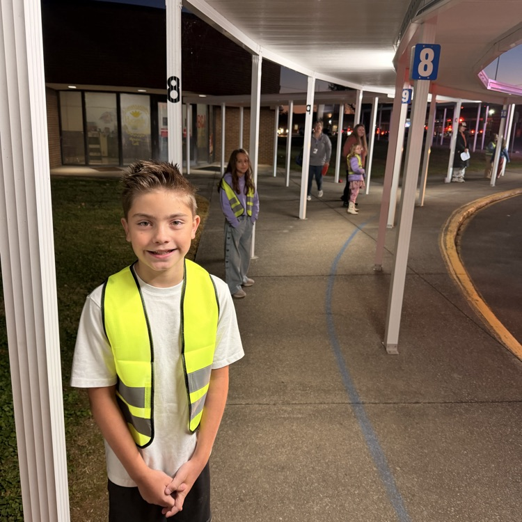 Child in yellow vest stands on sidewalk. Several people walk in distance, two in reflective vests.