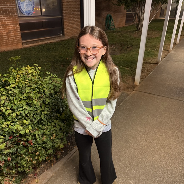 A young girl in a reflective vest stands on a sidewalk in front of a building.