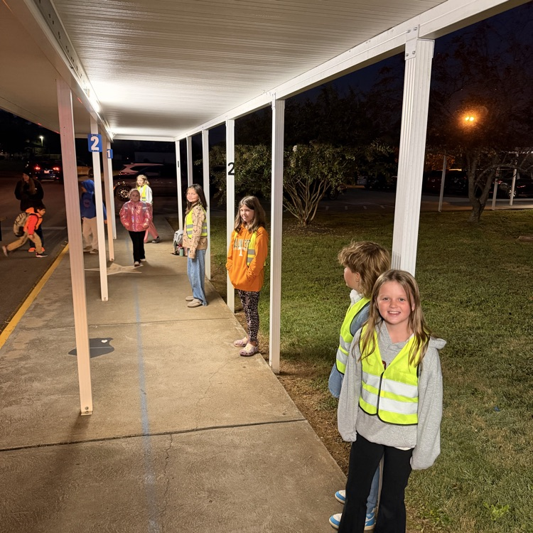 Students in reflective vests stand on sidewalk at school; some hold bags; one wears sneakers.