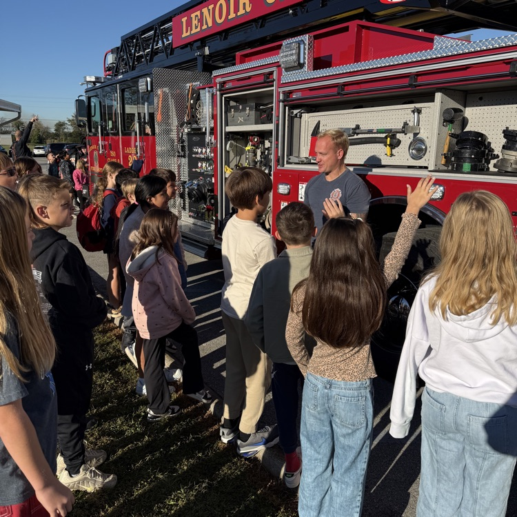 Children stand in line in front of a red fire truck with a man explaining something.