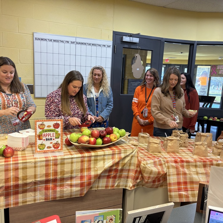 Women stand around tables with apples and paper bags; one person peels apples with a peeler.