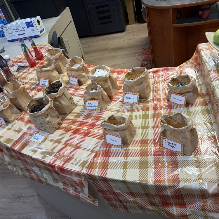 Table with brown bags, white labels, and small bottles of red and green sauce. Brown wooden floor.
