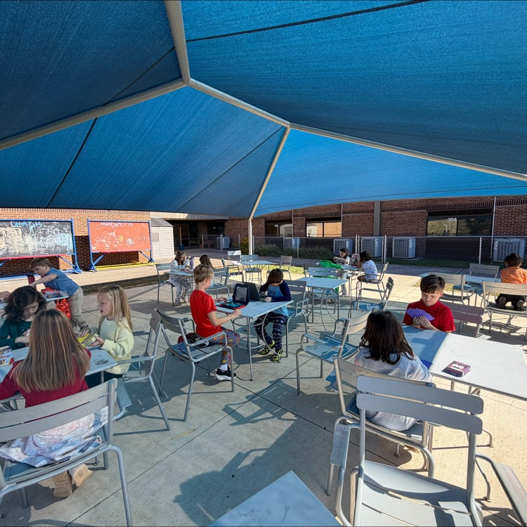 Students seated at tables under a blue canopy, working on their tablets and papers.