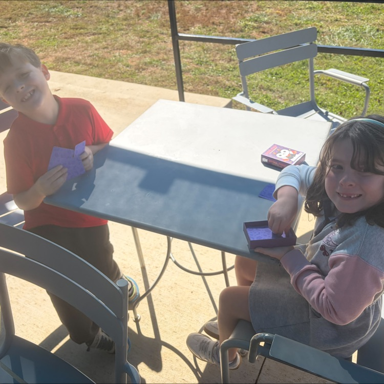 Two children seated at an outdoor table with papers and electronic devices in front of them.