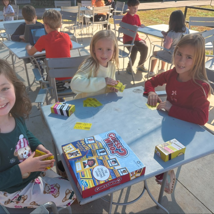 Three children sit at a table with a game board, yellow cubes, and other game pieces.
