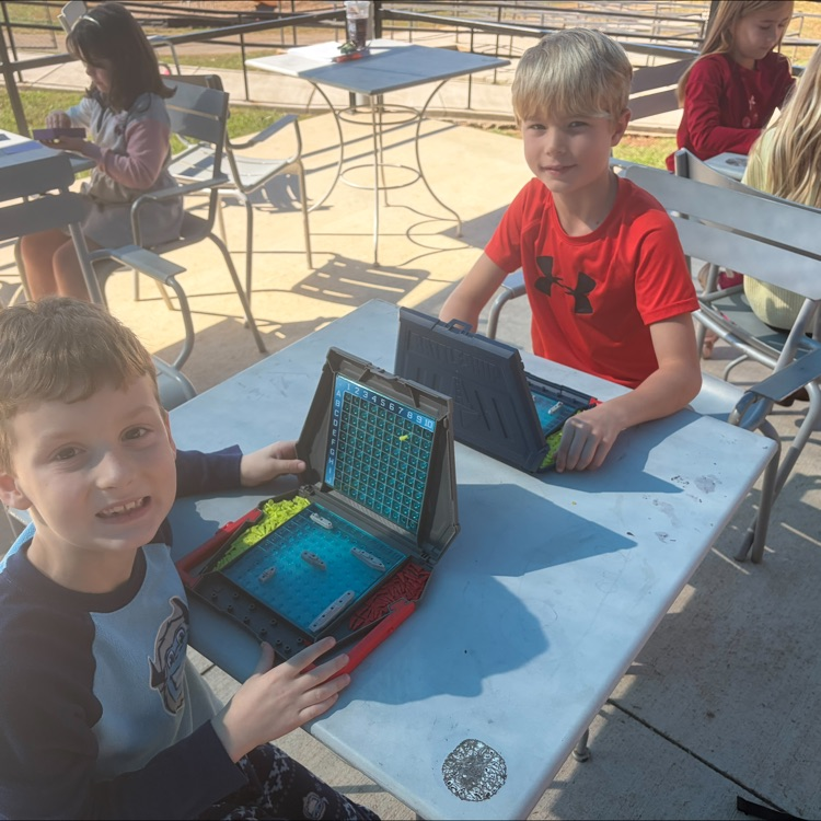 Two boys using electronic devices at a table in an outdoor seating area with other children.