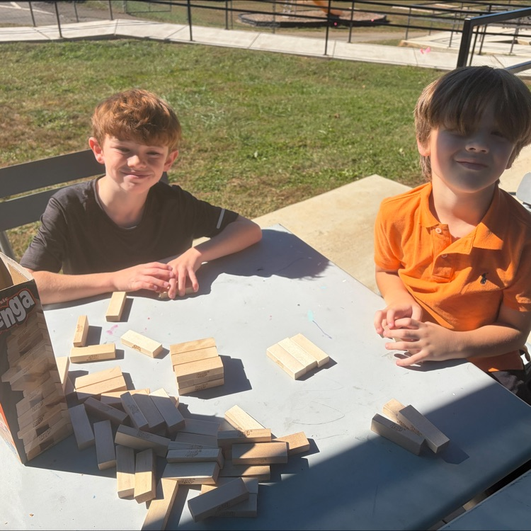 Two boys sit on chairs at a table with a box and blocks, smiling, on a sunny day.