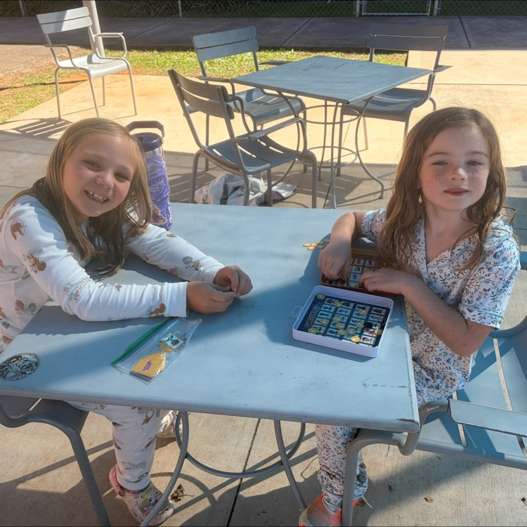Two girls sitting at a blue table with a tablet. One is holding a pencil.