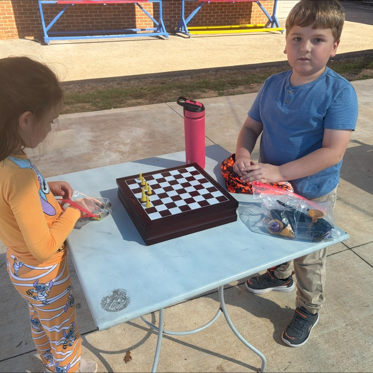Two children standing next to a table playing chess and holding plastic bags.