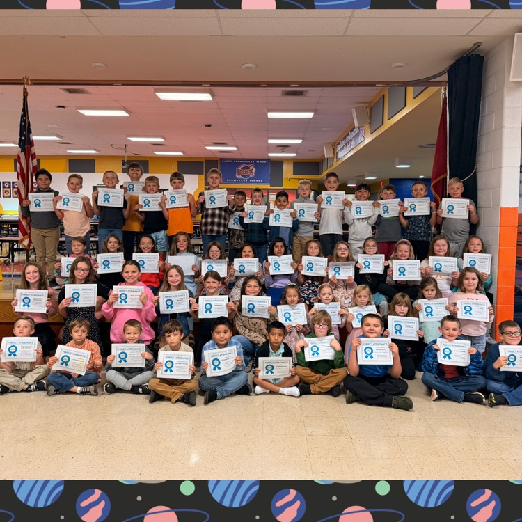 Classroom with students holding certificates; many standing and some sitting on floor, in front of flag and blue curtains.
