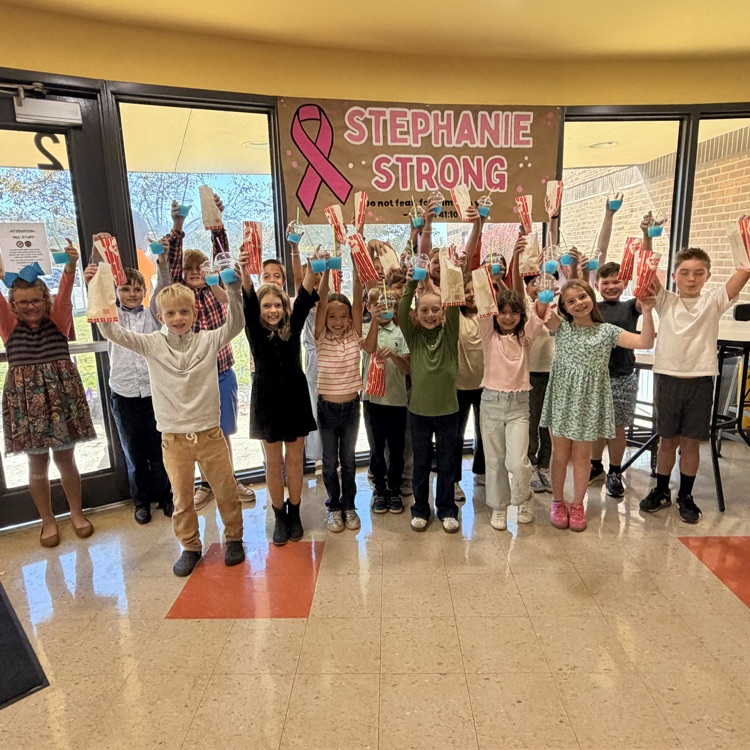 Group of children in a hallway holding up orange objects. Banner in background with pink ribbon and "STEPHANIE STRONG" text.