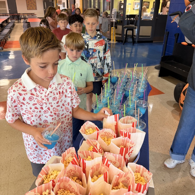 Four boys at a snack table with popcorn in paper bags, one holding a blue drink.