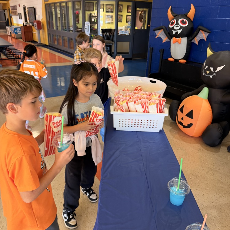 Children stand in line at a table with food and drinks, holding bags. Behind them are various Halloween decorations.