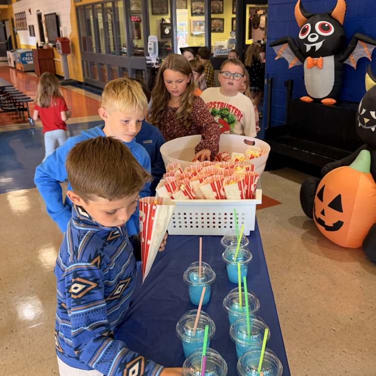 Children stand by a table with blue drinks and straws, a basket of popcorn, and a Halloween decoration.