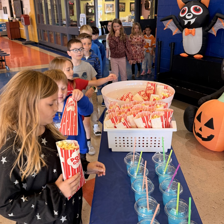 Children standing in line at a table with popcorn in buckets, cups with straws, and a pumpkin.