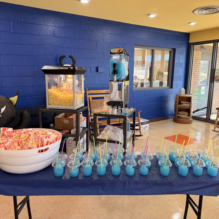 A blue room with a table full of blue drinks and straws. Bowl of popcorn and a popcorn machine.