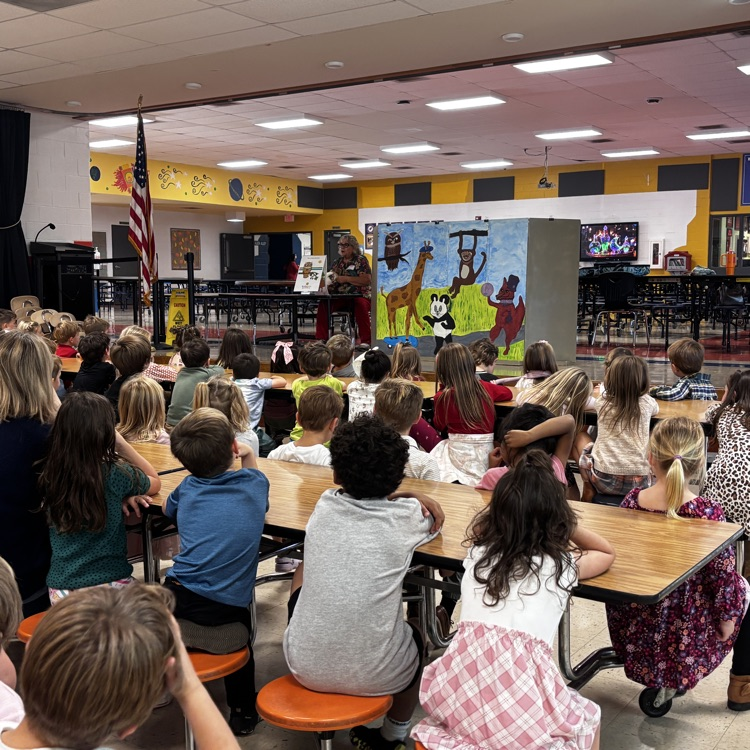 Children sit at tables in a classroom, listening to a person standing by a large poster.