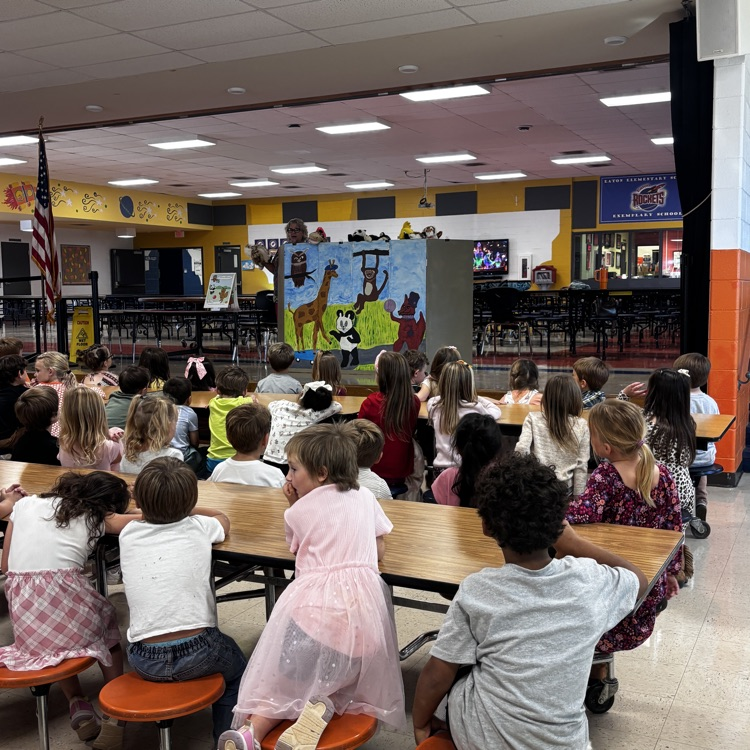 Many students seated at tables inside a room with colorful wall murals, listening to a presentation.