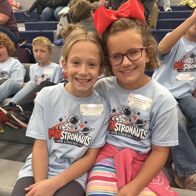 Two girls in matching shirts sit together, smiling for a photo. One wears a red bow; both have glasses. Behind them, more kids sit on bleachers.