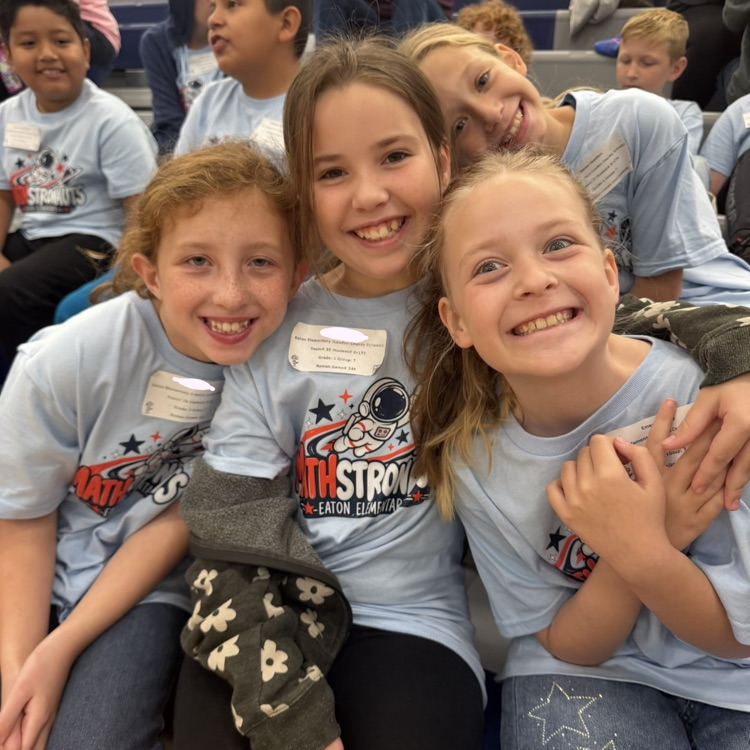 Four children wearing matching shirts smile and pose together in a school auditorium.