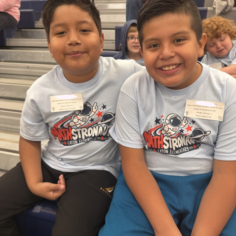Two smiling boys in matching shirts sit on a bleacher. Behind them, several other children sit on bleachers, some looking at phones.