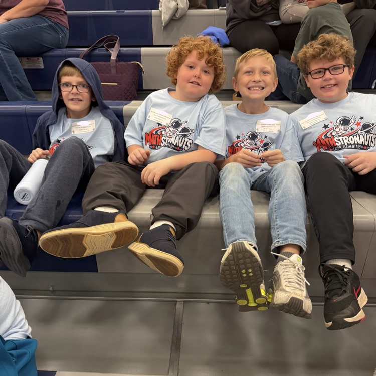 Four boys sit together on bleachers, wearing identical shirts with a logo. One has glasses, one wears a hoodie, and all wear sneakers.