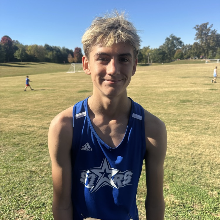 Boy in blue shirt with white stars, number 707, smiles in grassy field with distant runners.