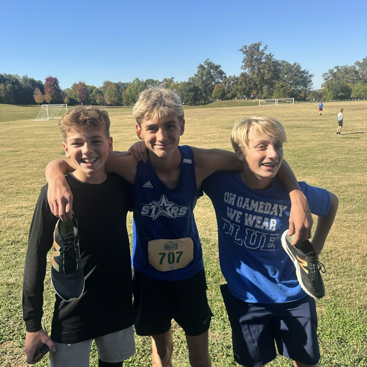 Three boys standing in a grassy field wearing athletic uniforms and holding shoes in their hands.