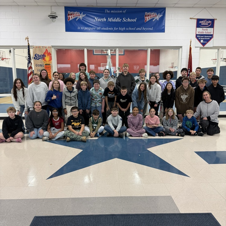 Large group of students in a school gym, standing and sitting, arranged around a star pattern.