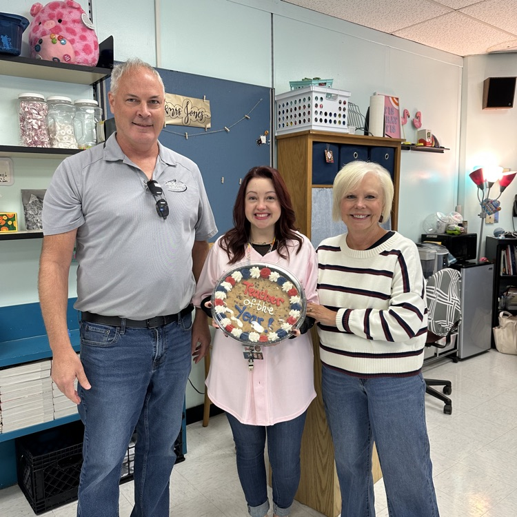 Three adults stand next to each other. A woman holds a circular object with a message on it.