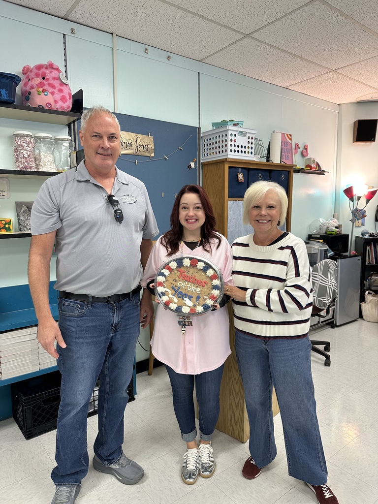 Three adults standing in a room with a young woman holding a circular item; shelves, boxes, and a plant are in the background.