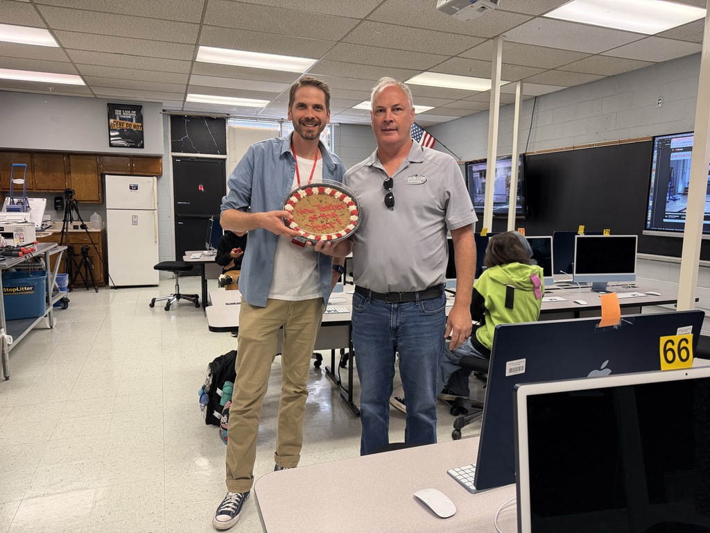 Two men in a classroom with computer equipment and a trophy. One man holds a trophy while the other wears a belt.