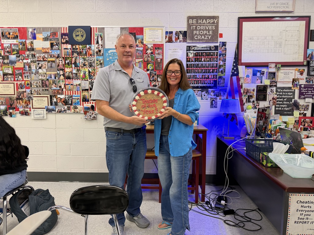A man and woman stand in a classroom, holding a circular plaque. The room has various pictures and papers on the wall.