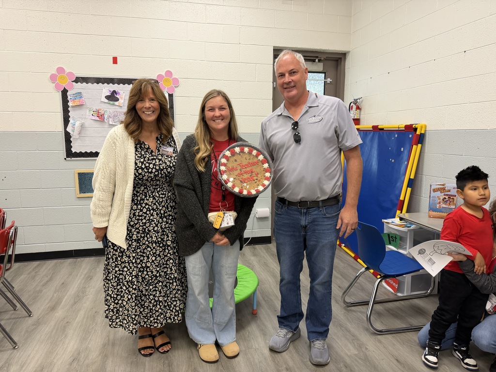 Four adults and two children in a classroom. One woman holds a trophy. One child holds a paper.