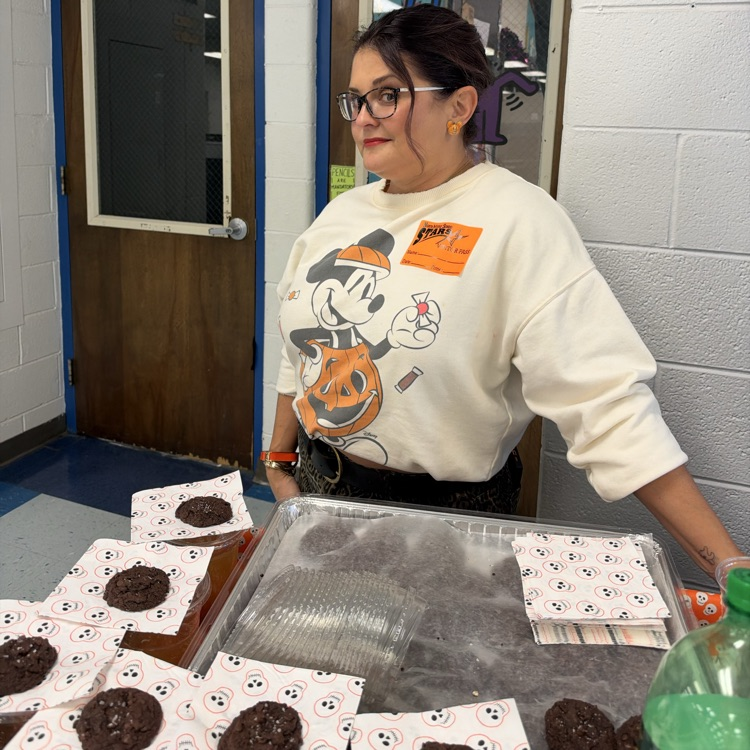 Woman wearing glasses and a Mickey Mouse sweatshirt standing behind a table with cookies on it.