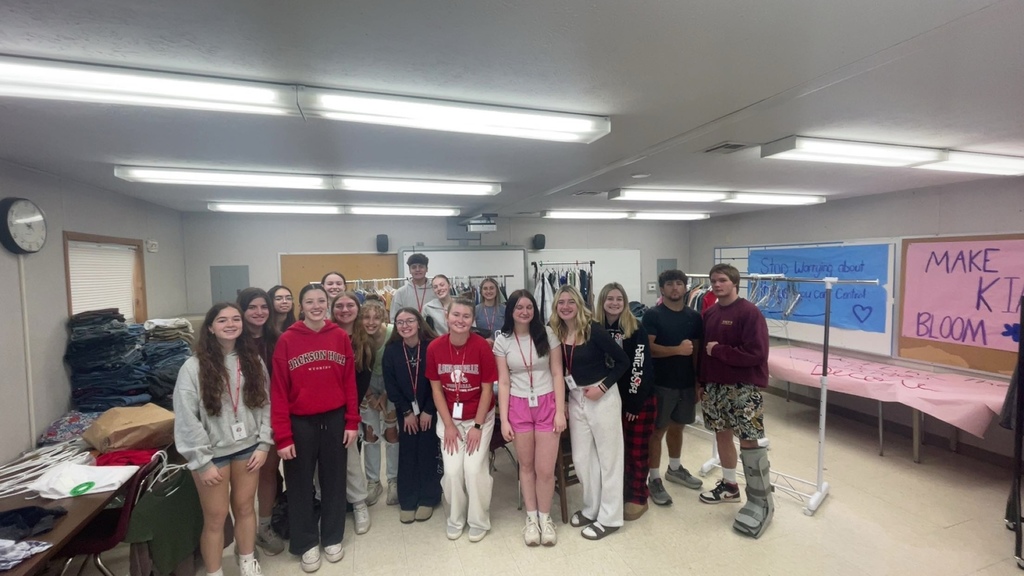 Group photo of students in a classroom with clothing donations behind them for the Squad Shop.
