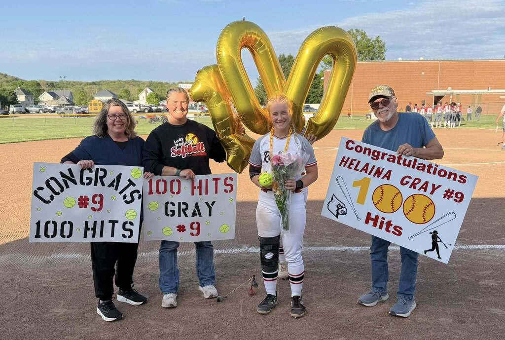 Student in softball uniform with family holding congratulatory signs for 100th hit.