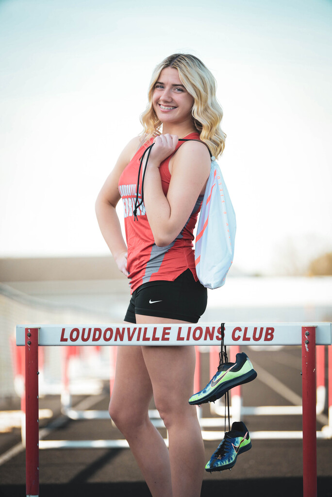 Photo of Cama on track standing behind a hurdle with her track bag and shoes.