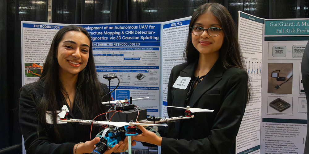 Two female students stand at a research poster session holding a quadcopter drone with a mounted camera, sensors, wiring, and propellers. Behind them are large academic posters with diagrams and text about autonomous UAV development, infrastructure mapping, and machine learning analysis. The scene appears to be an indoor conference or exhibition hall with display boards and technical visuals.