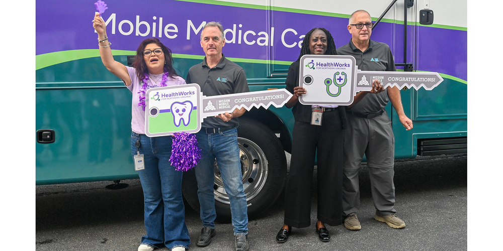 Four healthcare workers stand in front of a Mobile Medical Care van holding large congratulatory signs shaped like keys and medical icons.