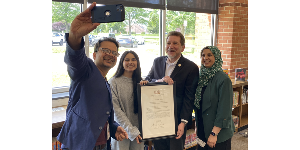 A group of people standing indoors in front of large windows and bookshelves. One person in the center holds a framed certificate or proclamation while another person holds up a smartphone to take a photo. Daylight and trees are visible outside the windows