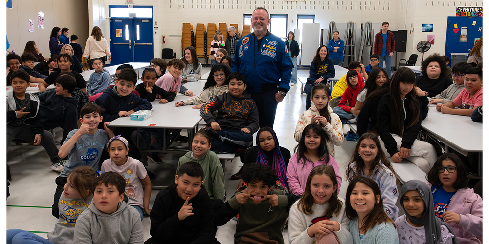 A person wearing a blue astronaut-style flight suit stands among rows of seated elementary-age students gathered around cafeteria tables in a school gym or multipurpose room, with teachers and school staff visible in the background.