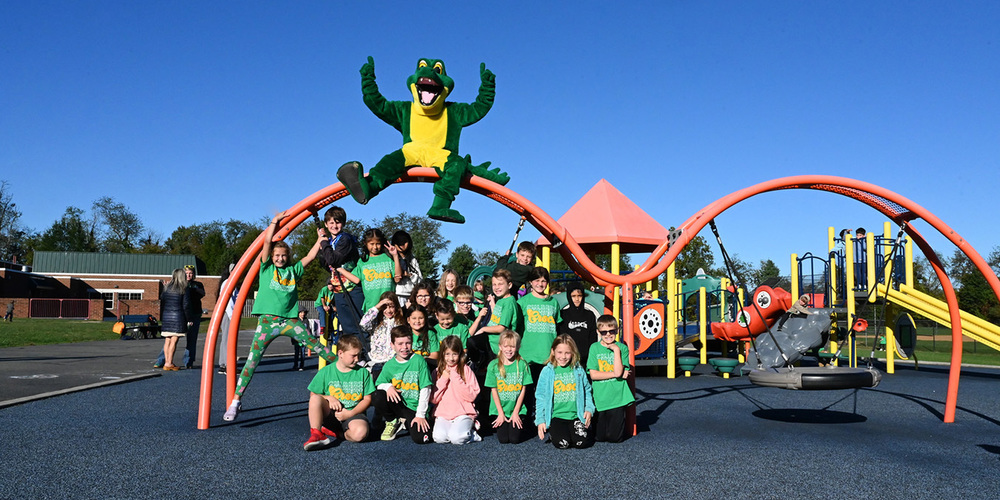 Group of children and adults wearing green shirts posing happily in front of a colorful playground with slides and climbing structures under a clear blue sky.
